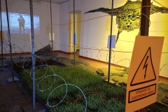Instalacija sa kukuruzom, pšenicom i sojom okružena bodljikavom žicom, električnom ogradom. Oko “vrta” je otpad , sa nadzorna kamera i animacija "Čuvar", UK Parobrod, Beograd, 2025.